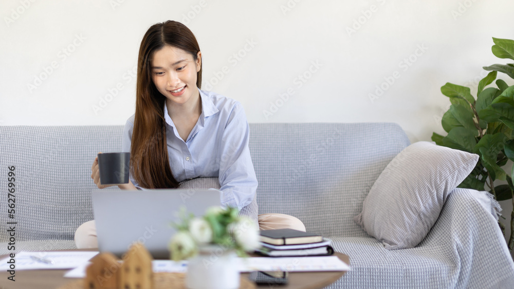 Beautiful Asian woman working on laptop and sipping coffee with smiling face in her home, Creating happiness at work with a smile, Freelancer working at home happily, Work from home. Beautiful Asian woman working on laptop and sipping coffee with smiling face in her home, Creating happiness at work with a smile, Freelancer working at home happily, Work from home.