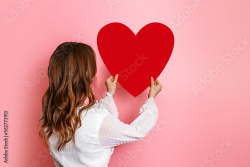 Girl hangs a big paper heart on a pink wall back view. Valentine's Day concept
