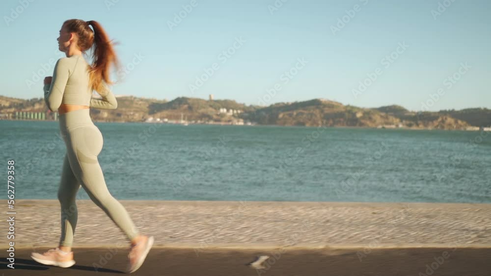 Confident fit female jogging by embankment area coastal city in beautiful sunny day do fitness training for cardio health. Tracking shot. Slow motion. Handheld. Athletic woman running, morning run Confident fit female jogging by embankment area coastal city in beautiful sunny day do fitness training for cardio health. Tracking shot. Slow motion. Handheld. Athletic woman running, morning run