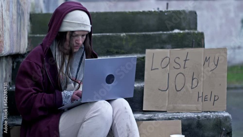 Homeless woman shivering in the cold with a laptop on her lap sits on cardboard amongst the rubbish. Next to her is a handwritten LOST MY JOB HELP poster. An unemployed IT professional.