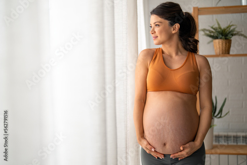 Beautiful pregnant woman touching her belly, standing by the window with the white curtains at home during the day. photo