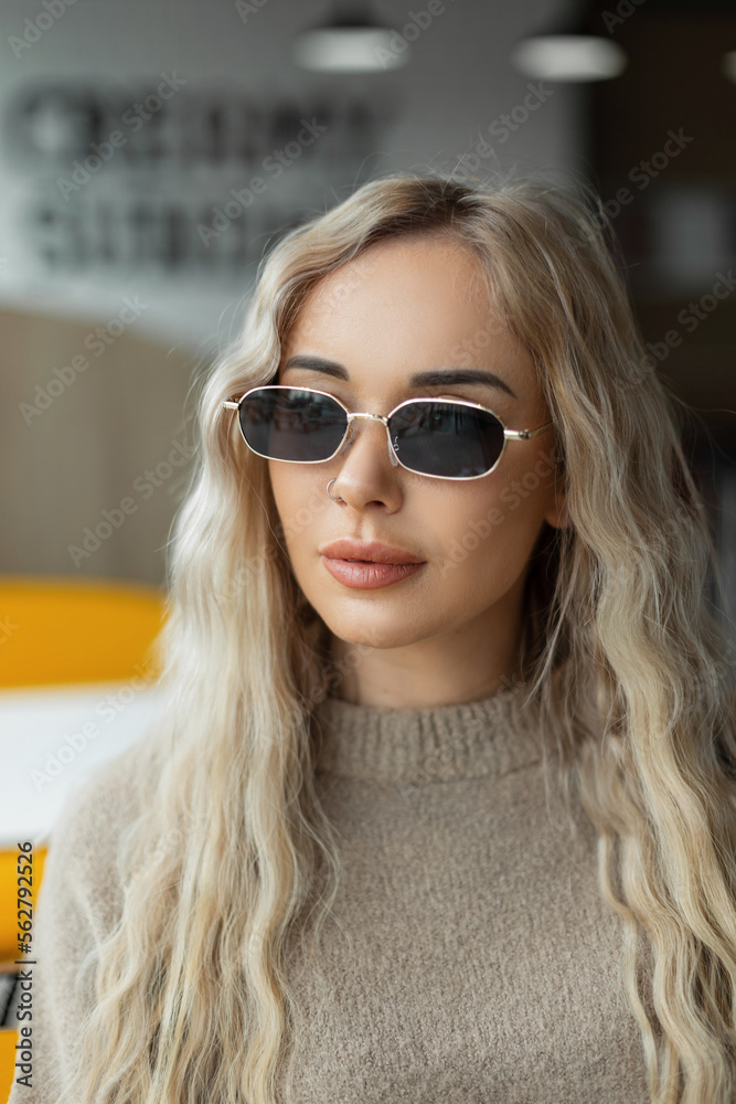 Naklejka premium Fashion female portrait of a beautiful young woman with vintage sunglasses with a curly blond hairstyle in a stylish beige sweater sits in a cafe