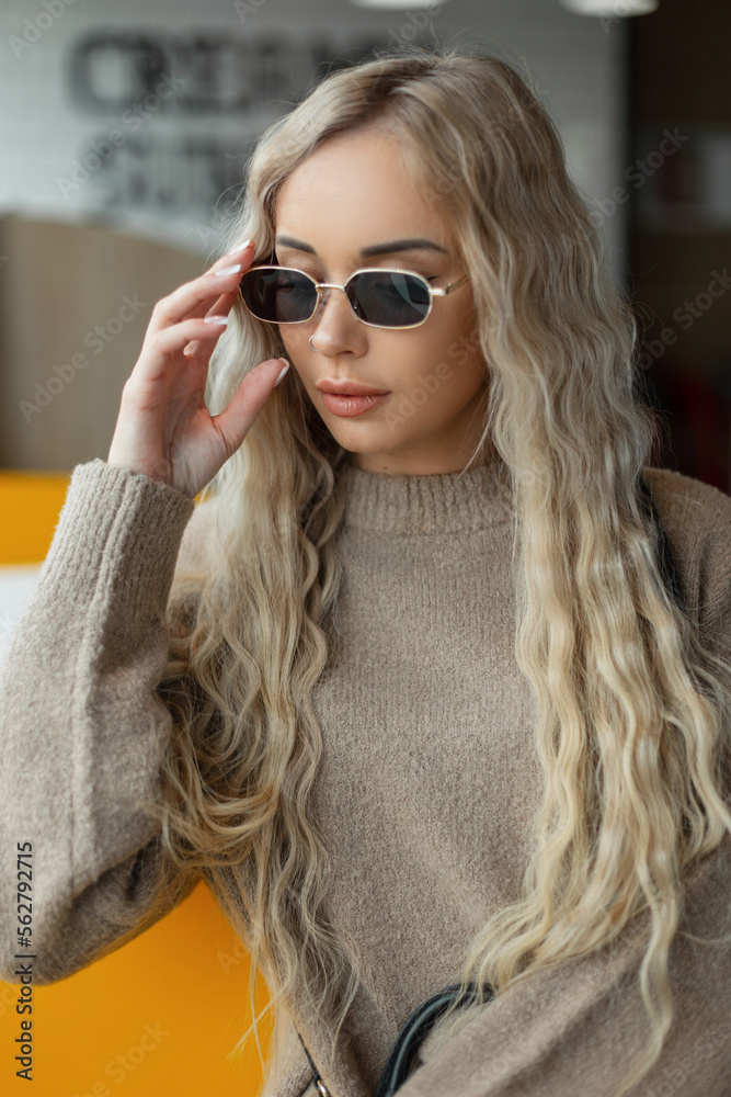 Naklejka premium Stylish beautiful cool blonde woman hipster with curly hairstyle in a beige sweater puts on vintage sunglasses and sits in a cafe