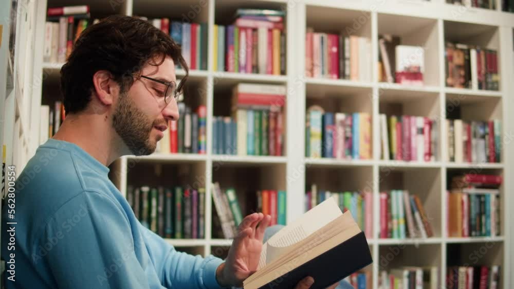 Man reading Book in University Library, Book Shop or Store, Spanish Student Wearing Glasses Standing near Bookshelf Reads Book and Exam Preparations. Man reading Book in University Library, Book Shop or Store, Spanish Student Wearing Glasses Standing near Bookshelf Reads Book and Exam Preparations.