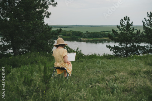 portrait of a young artist 10-13 years old in a yellow dress and a straw hat in nature painting a picture photo
