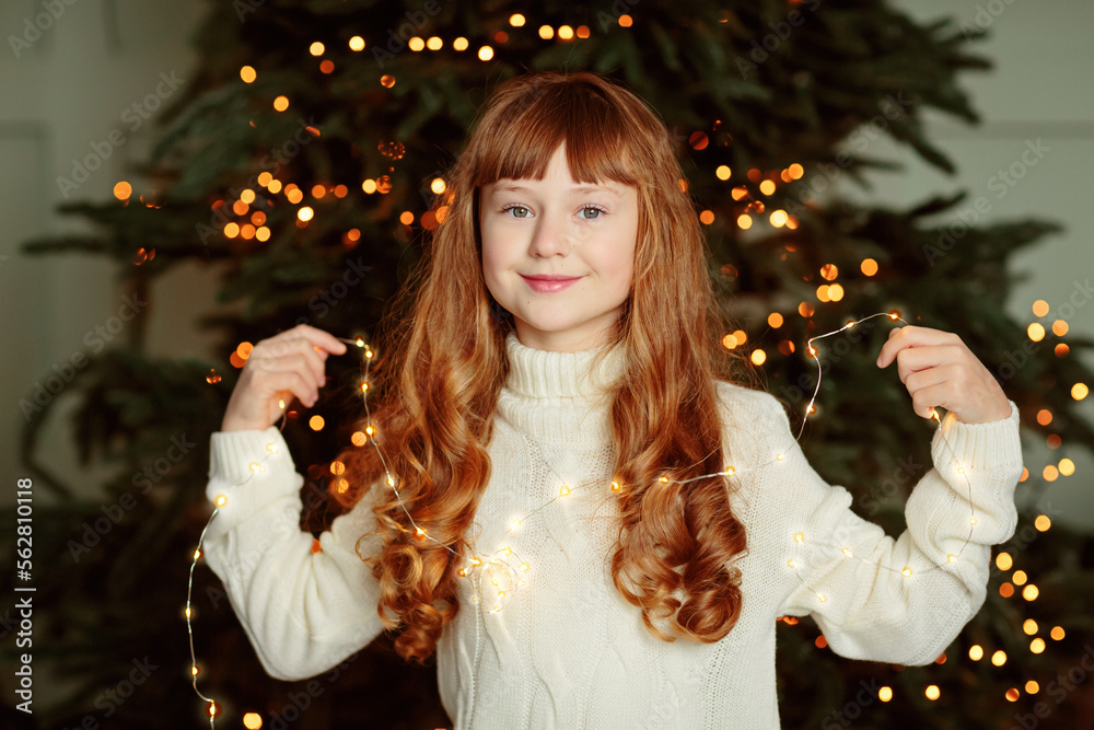 Happy young girl sitting by the Christmas tree in the cozy living room of the house and holding garland. Happy winter holidays concept