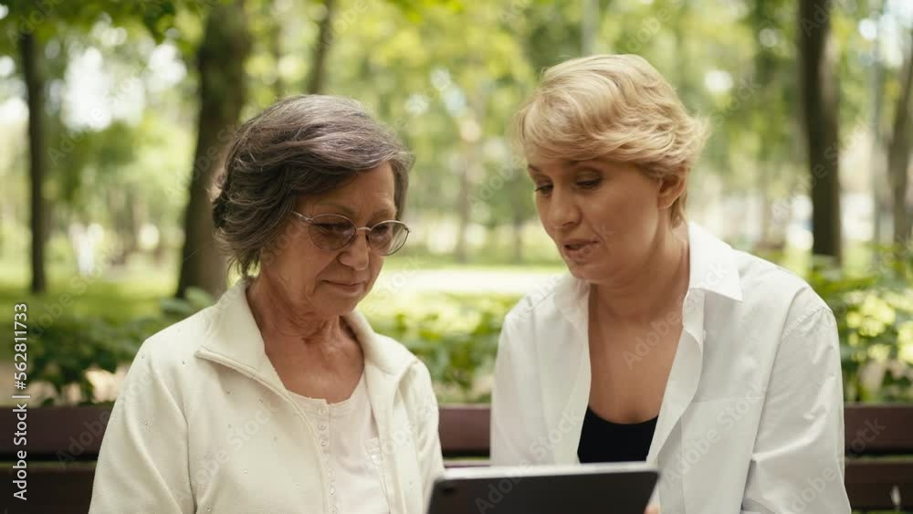 Daughter showing senior mother how to use a tablet while sitting on park bench Daughter showing senior mother how to use a tablet while sitting on park bench