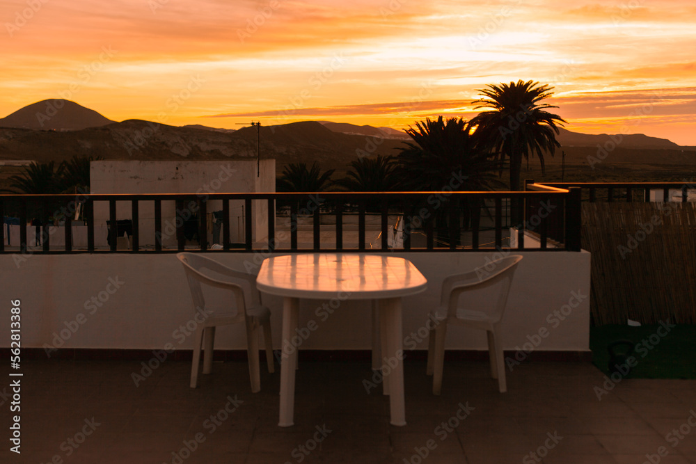 Terrace of a hotel  at sunset with view of mountains and silhouettes of palm trees. Table and chairs at a house terrace during the sunset or sunrise. Landscape of residential area.Real estate business