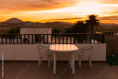 Terrace of a hotel at sunset with view of mountains and silhouettes of palm trees. Table and chairs at a house terrace during the sunset or sunrise. Landscape of residential area.Real estate business
