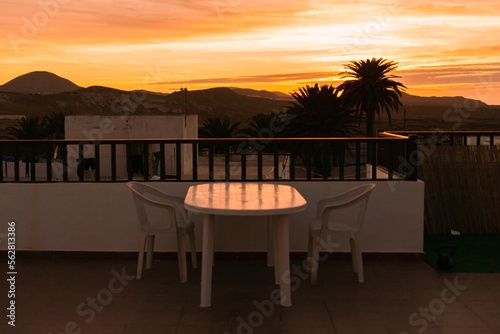 Terrace of a hotel at sunset with view of mountains and silhouettes of palm trees. Table and chairs at a house terrace during the sunset or sunrise. Landscape of residential area.Real estate business