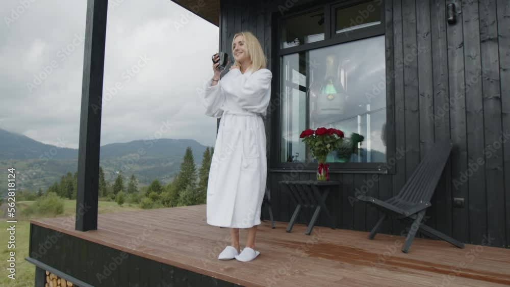 Happy female tourist enjoying her leisure time in countryside, mountainous corner. Caucasian, blonde haired woman in a white bath robe, drinking fresh morning coffee. High quality 4k footage Happy female tourist enjoying her leisure time in countryside, mountainous corner. Caucasian, blonde haired woman in a white bath robe, drinking fresh morning coffee. High quality 4k footage