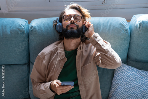 Young man wearing headphones listen podcast or music playing in smartphone app, happy guy relaxing holding using phone enjoy favorite songs relaxing sit on sofa at home. High quality photo