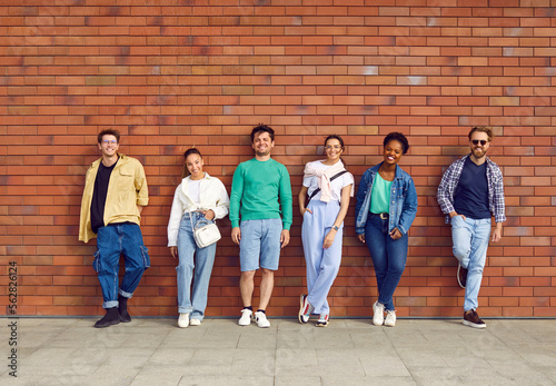Happy diverse friends. Six good looking young people in casual wear. Group portrait of handsome men and beautiful women in shirts, jumpers, jeans, sneakers and trainers leaning on brick wall in street