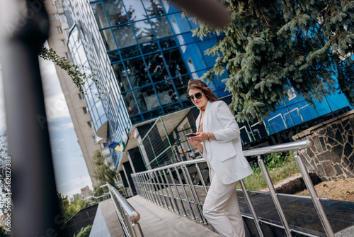 Pretty business woman in white suit and sunglasses using phone during break standing near modern office building with electric scooter in the foreground
