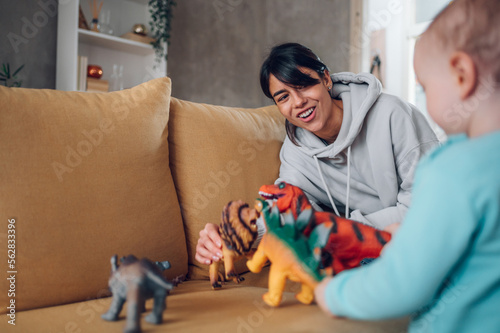 Mother and toddler son playing with dinosaurs toys at home on the couch