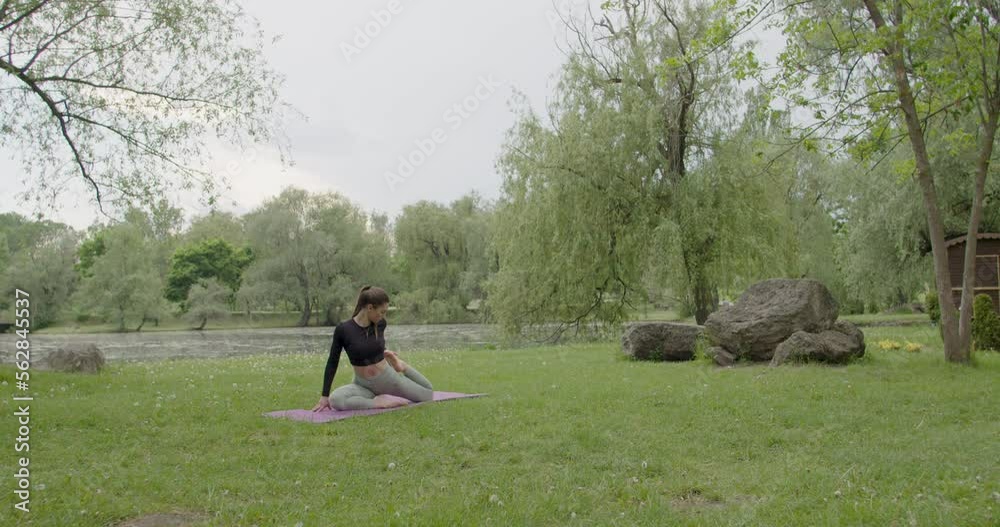 young woman, sitting on the grass in the park, prepares for meditation and does light exercises for relaxation. In sports clothes, next to a pond in the back and with instruments for meditation young woman, sitting on the grass in the park, prepares for meditation and does light exercises for relaxation. In sports clothes, next to a pond in the back and with instruments for meditation
