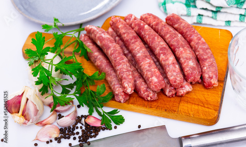 Pile of popular Spanish raw sausages longaniza with fresh greens, garlic and spices prepared for cooking on wooden cutting board with knife. Traditional meat products
