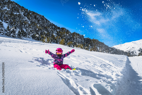 Smiling girl sitting on snow and throwing snow powder up to the air, mountains and forest in a background. Winter ski holidays, Andorra, Pyrenees