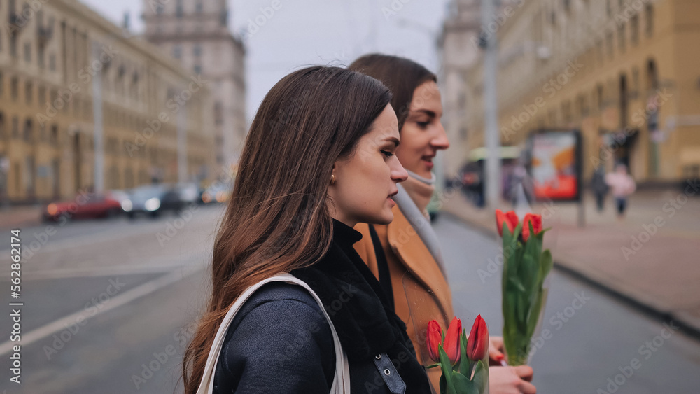 Two joyful friends in a jacket and coat are walking around the city with tulips. Two joyful friends in a jacket and coat are walking around the city with tulips.