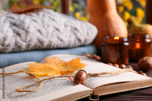 Book with autumn leaf as bookmark, acorns, scented candle and warm sweaters on wooden table, closeup