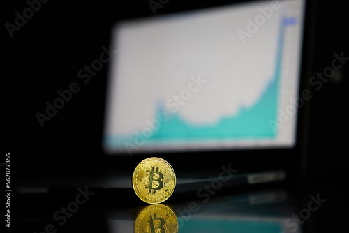 A single physical golden bitcoin in front of a screen of stock or cryptocurrency market candles or charts or graphs with a laptop
