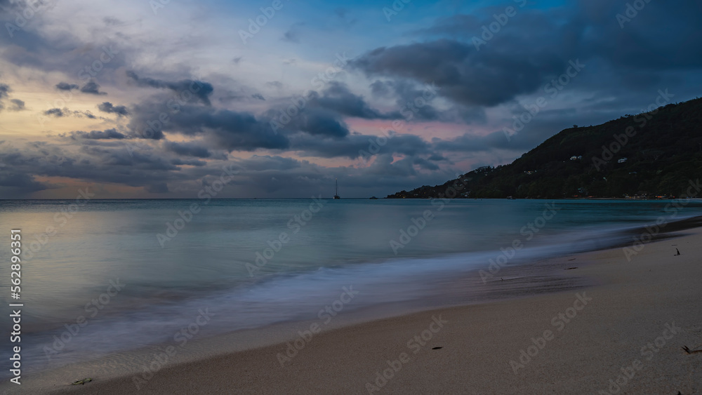 A quiet evening on a tropical island. The foam of turquoise ocean waves on a sandy beach. Clouds in the sky, highlighted in pink, orange. The yacht is on the horizon. Seychelles. Mahe. Beau Vallon