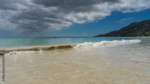 A long wave rolls onto the shore, twisting. Turquoise water mixes with the sand of the beach. Picturesque clouds in the blue sky. A hill in the distance. Seychelles. Mahe.Beau Vallon