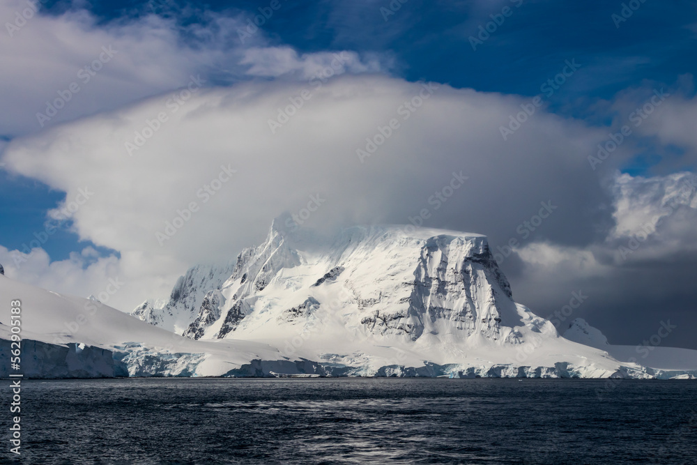 Mountain peak on Antarctic Peninsula. Ocean in foreground. Cloudy blue sky above. Cloud sitting on top of peak. 
