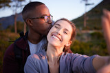 Interracial couple, kiss and smile for selfie, travel or love for adventure, journey or hiking together in nature. Happy man kissing woman smiling in happiness for photo moments, trip or traveling