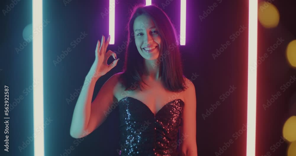 Portrait of satisfied young lady showing OK hand gesture and smiling standing on beautiful black background with neon lamps and bokeh lights Portrait of satisfied young lady showing OK hand gesture and smiling standing on beautiful black background with neon lamps and bokeh lights