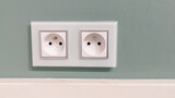 two white french european electrical outlets with plug inserted into modern neutral green wall with copy space banner