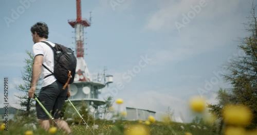 Hiker walking with hiking poles past the camera in foreground are yellow flowers and in the backgroun is radio tower.