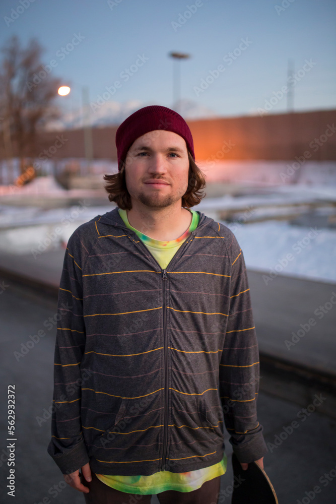 A young skateboarder stands in front of a skate park.