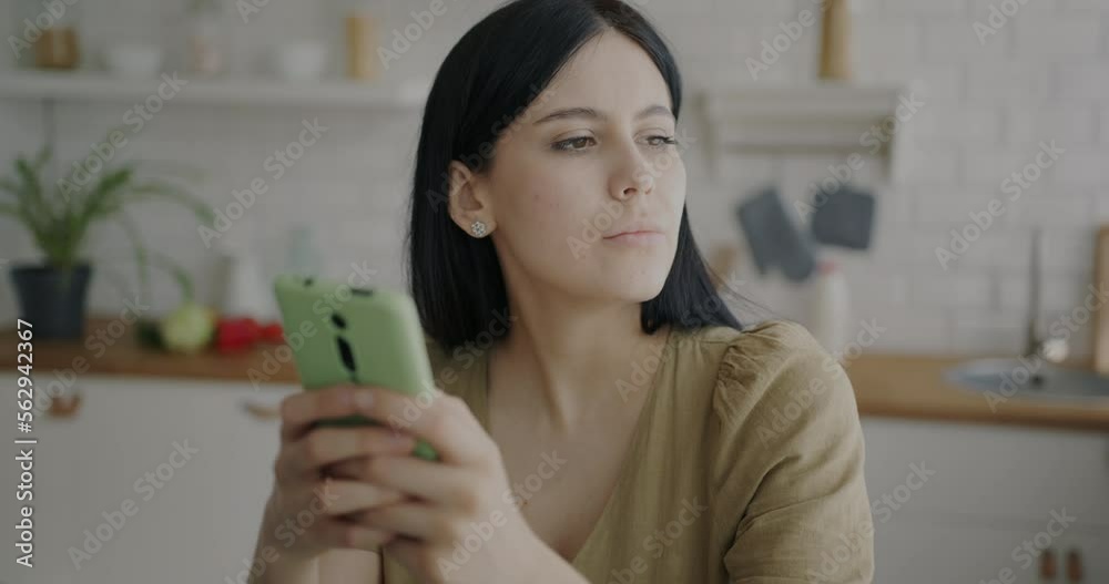 Portrait of pensive young lady using smartphone texting looking at display indoors in kitchen. Online communication and gadget concept. Portrait of pensive young lady using smartphone texting looking at display indoors in kitchen. Online communication and gadget concept.