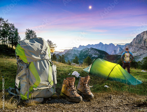 Hiker man standing next to Hiking Equipment, Tent, Backpack and Boots under a moon night sky at amazing twilight hour. Alps, Triglav National Park, Slovenia. photo