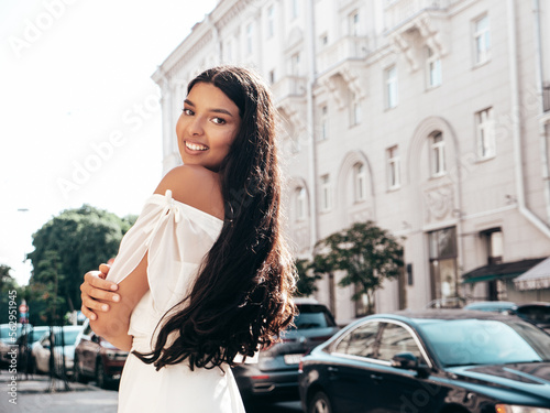 Young beautiful smiling hipster woman in white dress. Sexy carefree model posing on the street background at sunset. Positive female outdoors at warm sunny day. Cheerful and happy. Turns around