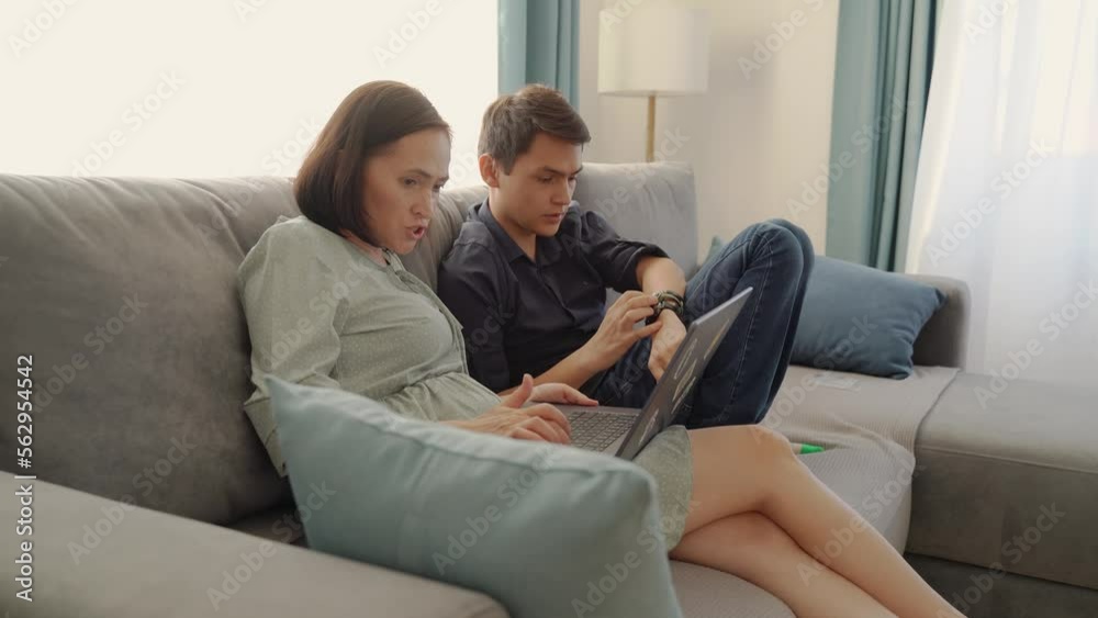 A woman in a dress and an adult son are sitting on the couch with a laptop. Couple sit on couch using laptop buy goods services on internet, enjoy easy comfort e-banking app. A woman in a dress and an adult son are sitting on the couch with a laptop. Couple sit on couch using laptop buy goods services on internet, enjoy easy comfort e-banking app.
