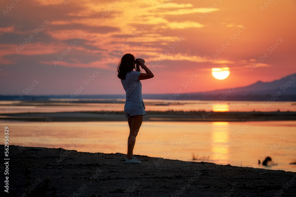 On safari in Zimbabwe: Silhouette of a Fit Woman standing on the Banks of the Zambezi River, observing African nature through binoculars, against red setting sun reflecting on water surface.