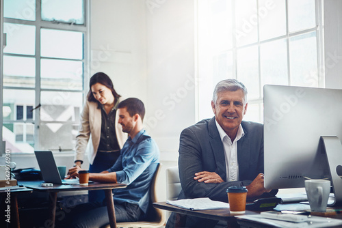 Senior, portrait and business man by computer in office workplace. Pc, coworking and happy elderly male employee working on marketing report, advertising or sales project with colleagues in company.
