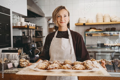 Photography Welcoming female baker holding freshly baked almond croissants in  background of bakery shop
