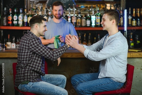Two young men in casual clothes are smiling and talking while sitting at bar counter in pub