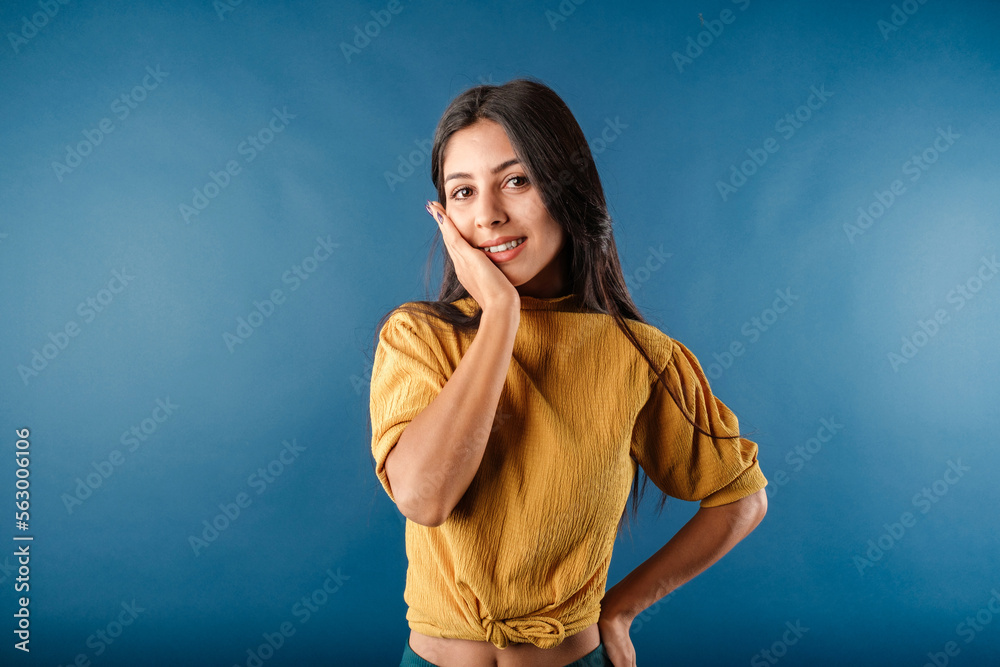 Young dark-haired woman wearing yellow top isolated over blue background looks at the camera laughing while touching cheek with the palm of one hand. Very pleased and impressed. Young dark-haired woman wearing yellow top isolated over blue background looks at the camera laughing while touching cheek with the palm of one hand. Very pleased and impressed.