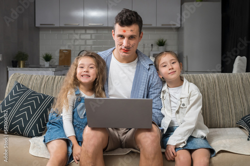 Junior schoolgirl paints father lips and enjoys playing with sister, looking at camera. Father with exhausted expression tries to work on laptop getting distracted at home