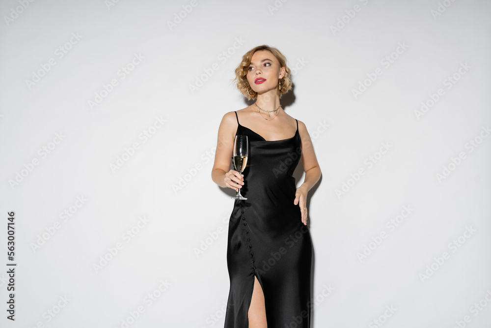 blonde woman in satin slip dress holding glass of champagne while standing with hand on hip isolated on grey. blonde woman in satin slip dress holding glass of champagne while standing with hand on hip isolated on grey.