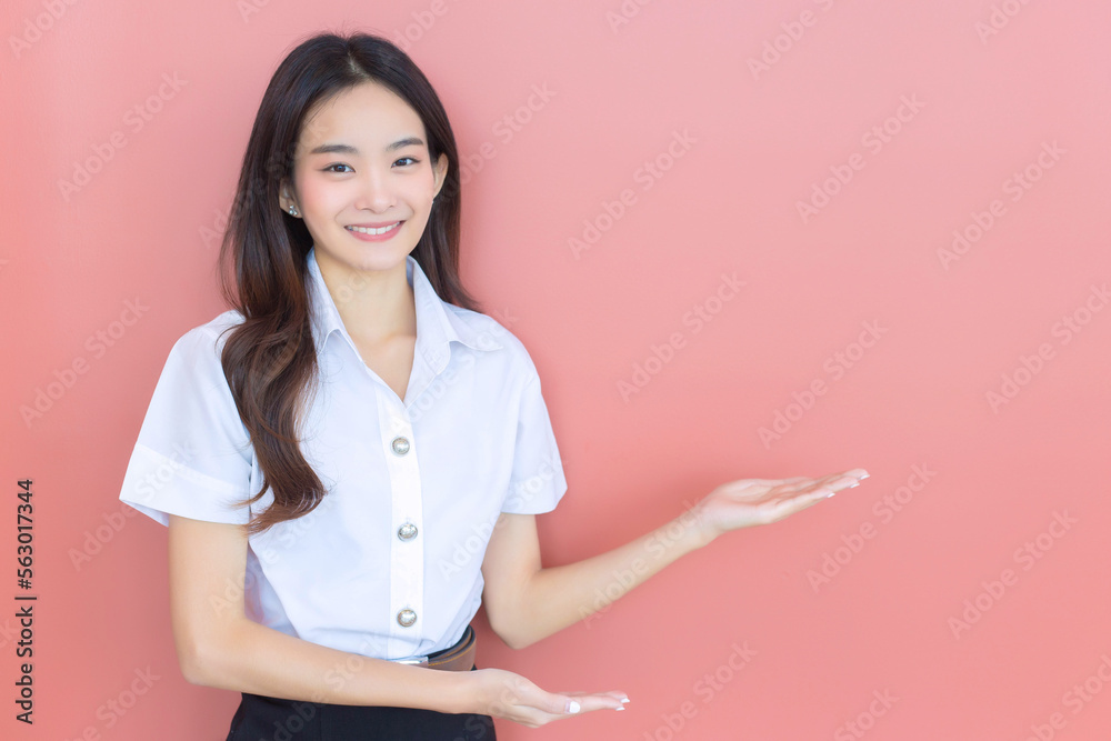 Portrait of adult Thai student. Beautiful Asian young woman student in uniform is smiling and looking at camera to present something confidently on pink background in the university Portrait of adult Thai student. Beautiful Asian young woman student in uniform is smiling and looking at camera to present something confidently on pink background in the university