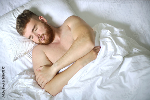 Handsome young calm sleepy bearded man with beard is sleeping well on side in bed in bedroom on pillow with his eyes closed in the morning  resting. Good healthy dream  rest. White linens. Top view.