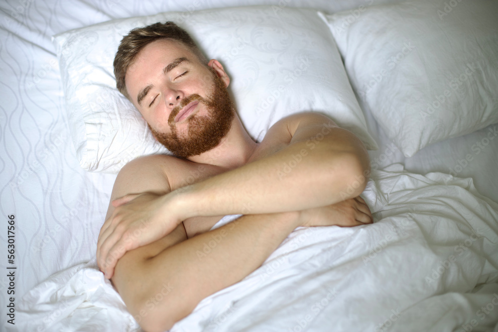 Handsome young calm sleepy bearded man with beard is sleeping well on side in bed in bedroom on pillow with his eyes closed in the morning, resting. Good healthy dream, rest. White linens. Top view. Handsome young calm sleepy bearded man with beard is sleeping well on side in bed in bedroom on pillow with his eyes closed in the morning, resting. Good healthy dream, rest. White linens. Top view.