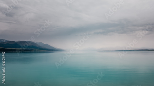 Clouds and mist hang over over the turquoise blue water of Lake Tekapo in New Zealand with snow capped mountains in the distance