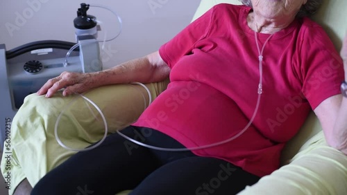 Person sit on chair using Oxygen breathing machine at home. old woman in red t-shirt. Breathing device for asthma and coronavirus. senior have problem with lungs.