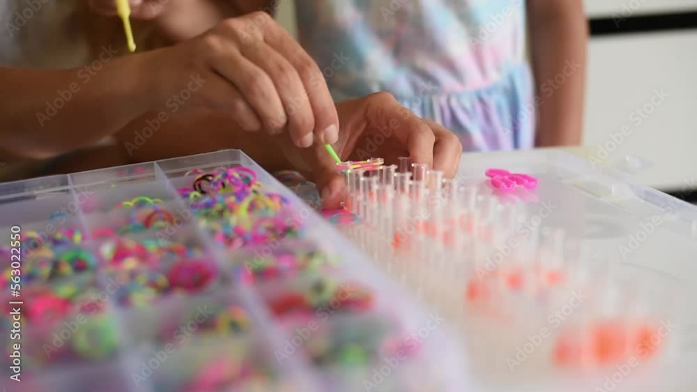 Mother's hands show girl how makes accessories bracelet from silicone rubber bands. Many different colors rings in box. Make necklace by hook. Hobby for children, motor skills. gift for friend